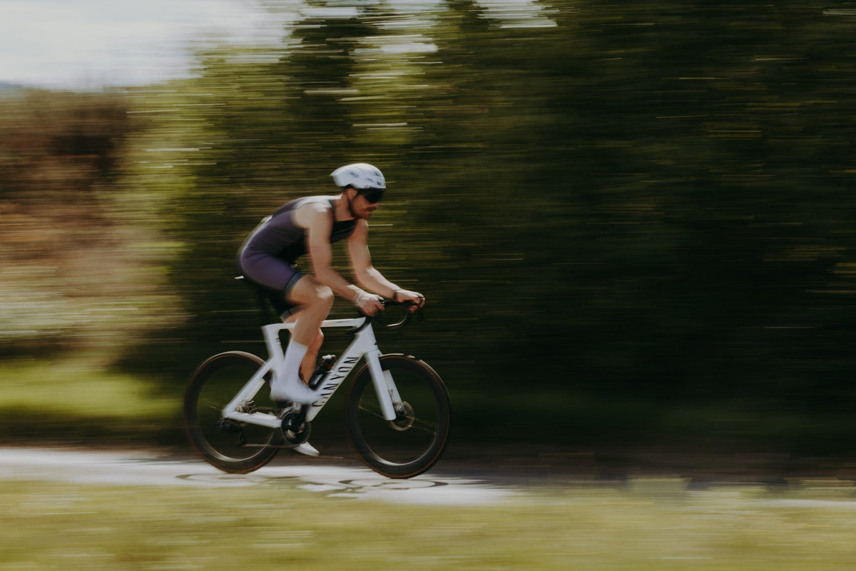 Male triathlete riding a road bike at speed while wearing a dark one-piece triathlon suit, with motion blur capturing the pace and performance of outdoor cycling training.