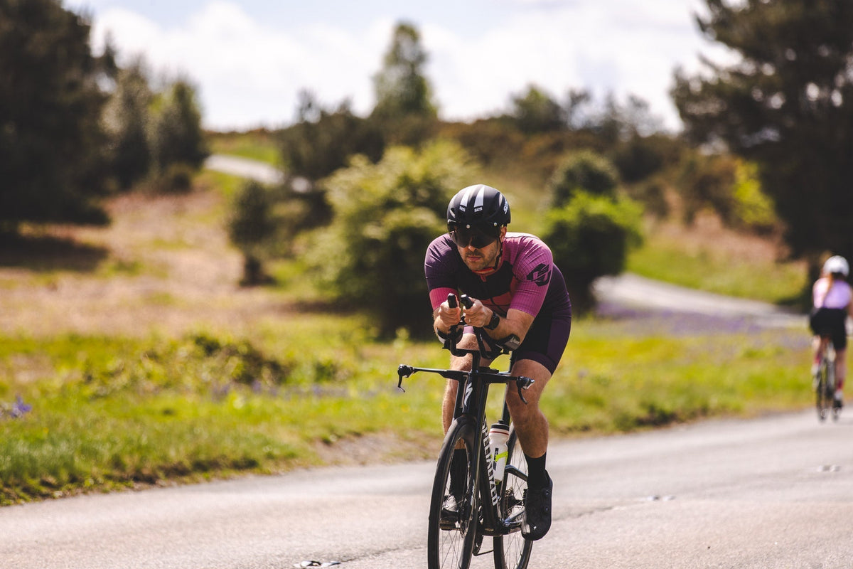 Cyclist wearing a purple and pink SLS3 triathlon suit, helmet, and sunglasses while riding a road bike outdoors surrounded by lush green vegetation.