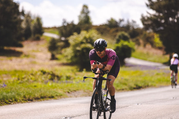Cyclist wearing a purple and pink SLS3 triathlon suit, helmet, and sunglasses while riding a road bike outdoors surrounded by lush green vegetation.