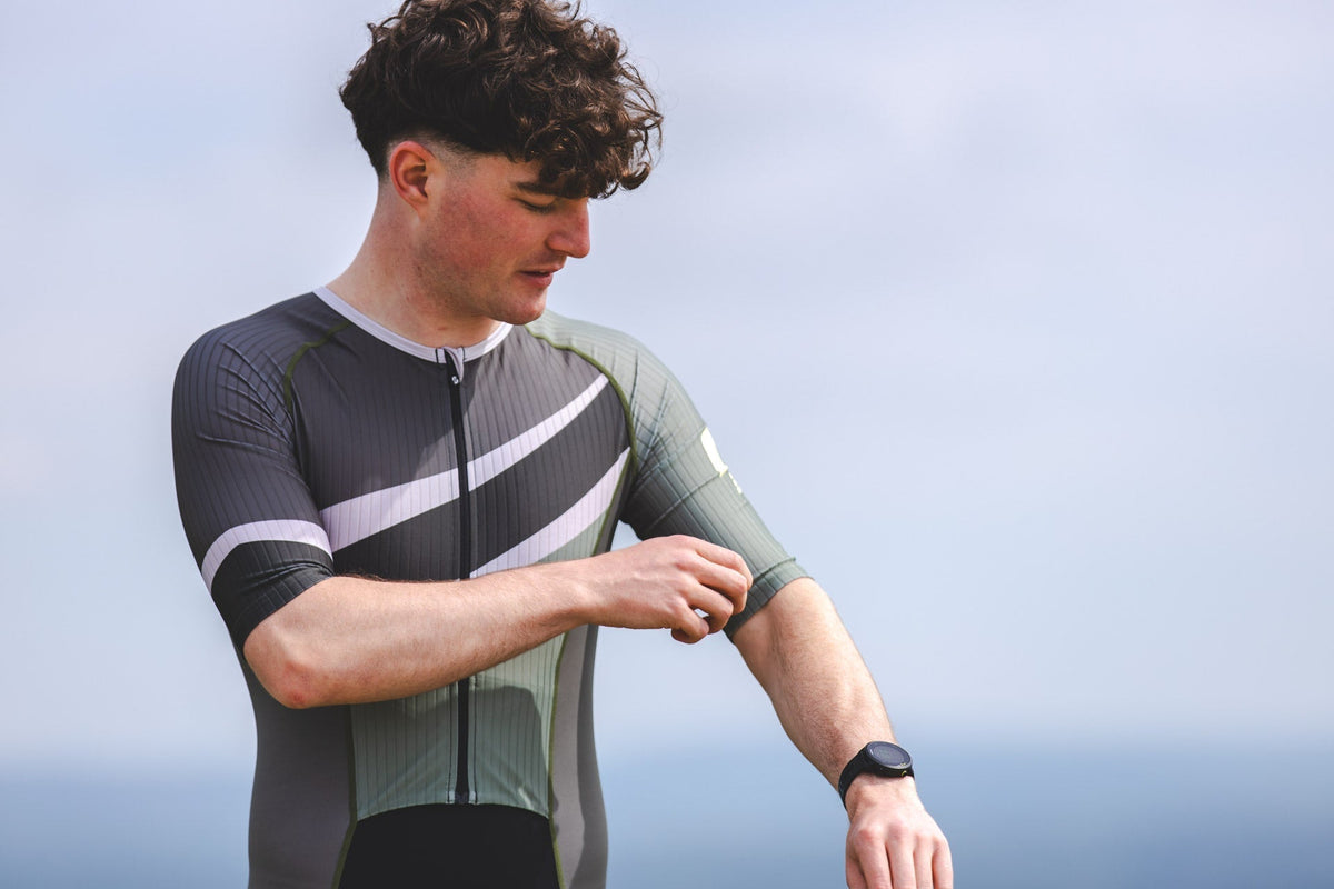 Young male triathlete adjusting the sleeve of his olive green SLS3 triathlon suit before training, showcasing the close-fitting fabric and aerodynamic design against a coastal backdrop.