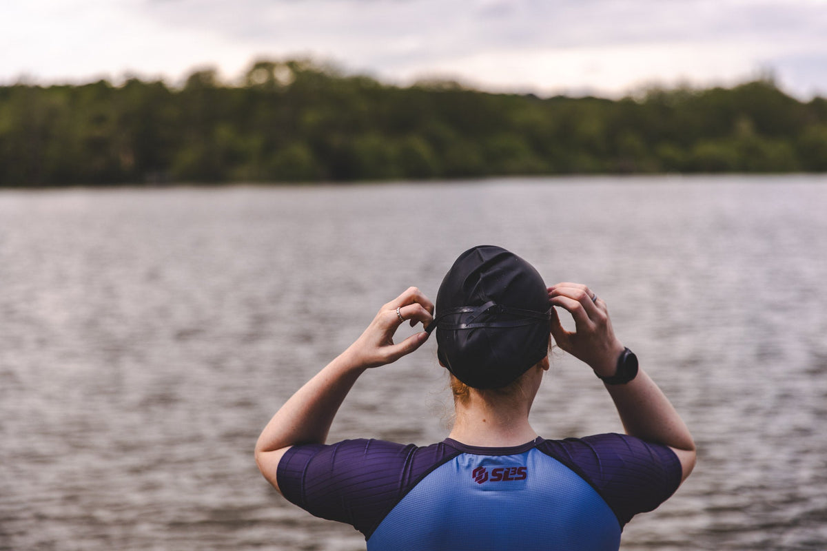 Female triathlete in a blue and purple SLS3 tri suit looking at a body of water before a swim, wearing a swim cap.