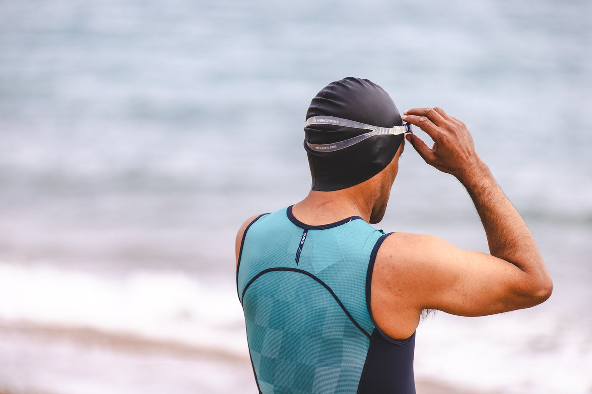 Triathlete wearing a teal and black SLS3 tri suit, adjusting swimming goggles while wearing a black swim cap outdoors by the water.