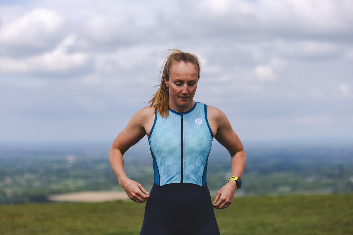 A female triathlete wearing a light blue and black SLS3 tri suit standing outdoors with a scenic mountain landscape in the background.